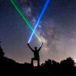 A silhoutte of a man standing on a hill top in the dark with two neon flash lights in his hand pointing the beam towards the sky