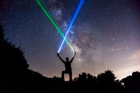 A silhoutte of a man standing on a hill top in the dark with two neon flash lights in his hand pointing the beam towards the sky